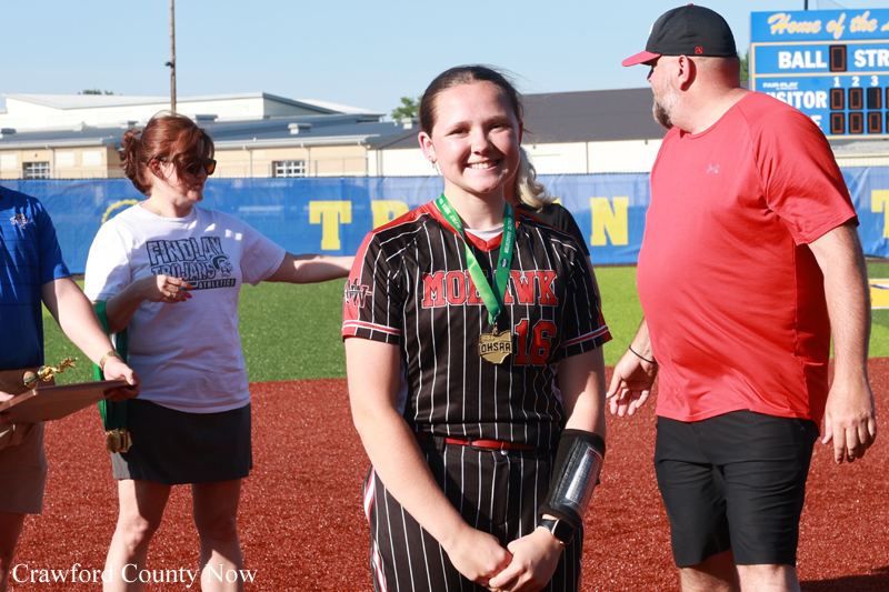 Smiling young softball player in a black pinstriped uniform with a medal around her neck, standing on the field as teammates and a coach congratulate her on a sunny day.