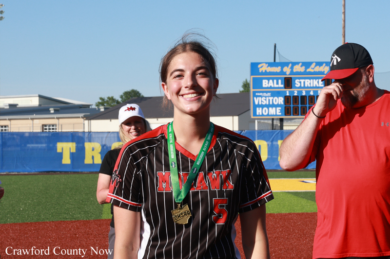 Smiling female softball player in a black pinstripe jersey with number 5 and a gold medal around her neck on a softball field.