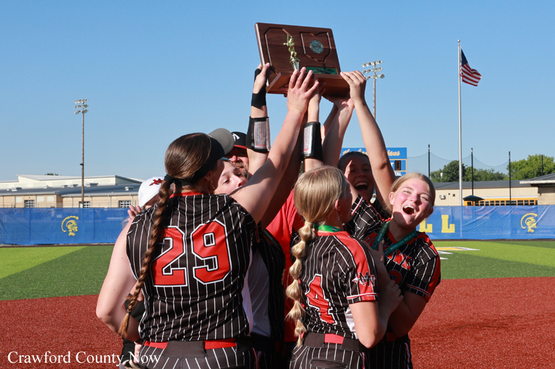 Girls' softball team in black pinstripe uniforms celebrate on a baseball field, lifting a trophy together.