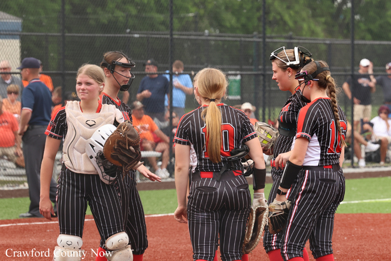 Girls' softball team in black pinstripe uniforms huddle on the infield, catcher with helmet, after a game on a dirt field.