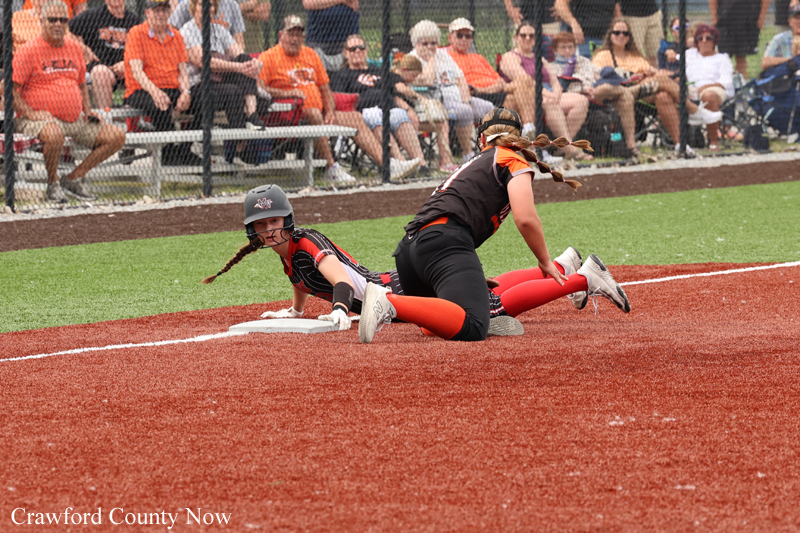 Softball players in black and orange collide at home plate as one slides in while a teammate reaches toward the plate; spectators watch from the stands.