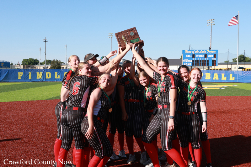 Girls' softball team in black pinstripe uniforms lift a trophy on a softball field, celebrating a win with smiles and raised arms on red infield dirt.