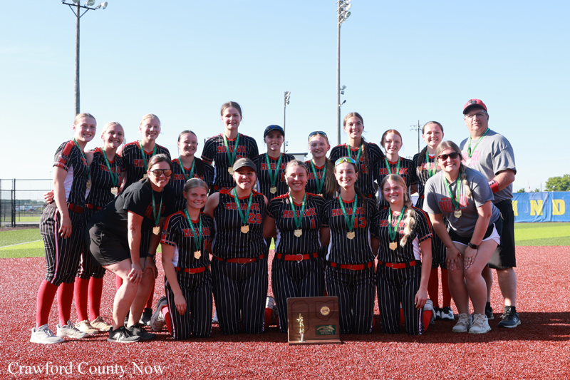 Softball team posing on a field after a win, wearing striped uniforms with red belts and medals, a plaque/trophy in front center.