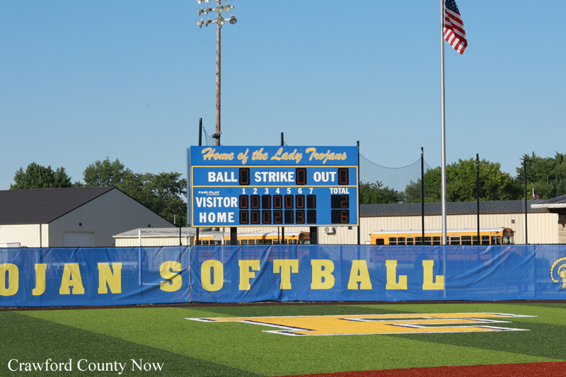 Scoreboard at a blue softball field banner reading 'SOFTBALL' with 'Visitor' and 'Home' scores visible in the stands area. American flag in the background.