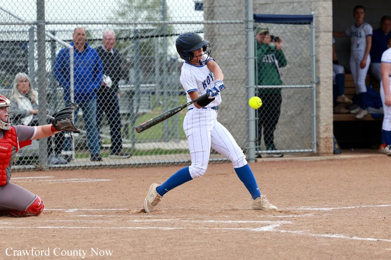 A young female softball batter in a white uniform swings at a pitch while the catcher in red gear watches from the left, with spectators behind a chain-link fence.