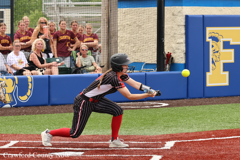 Female softball player in a black pinstripe uniform and red socks swings at a pitch near home plate, with a crowd watching behind a fence.