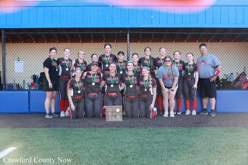 Girls' softball team in black pinstripe uniforms with medals, posing with coaches and a trophy in front of the dugout.