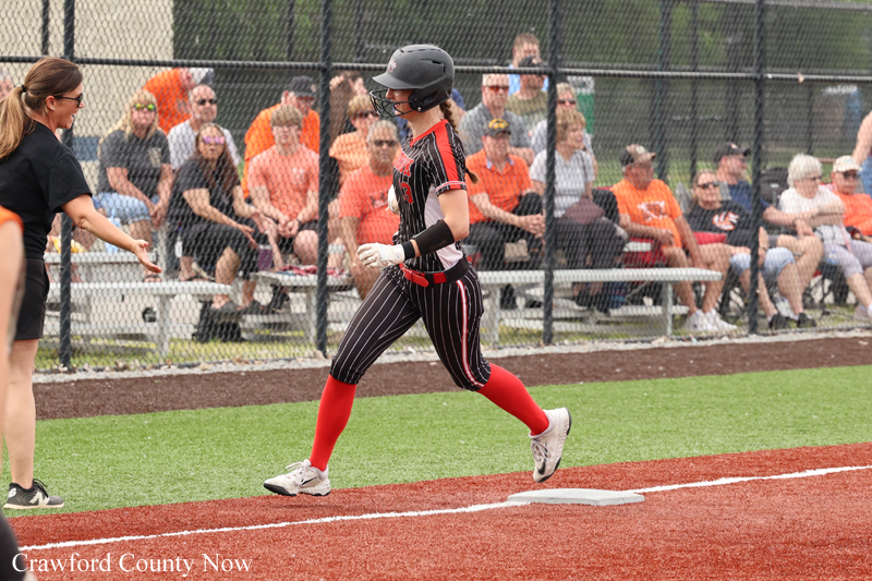 Softball player in a black pinstripe uniform runs toward home plate with helmet on as spectators watch from behind a chain-link fence.