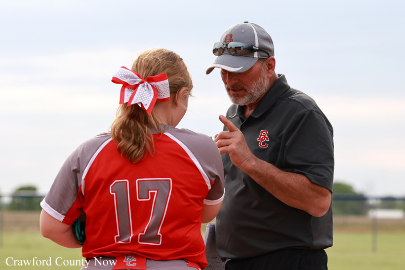 Coach in a dark polo talks to a young player in a red jersey with number 17 on a softball field, gesturing with his finger as he explains.
