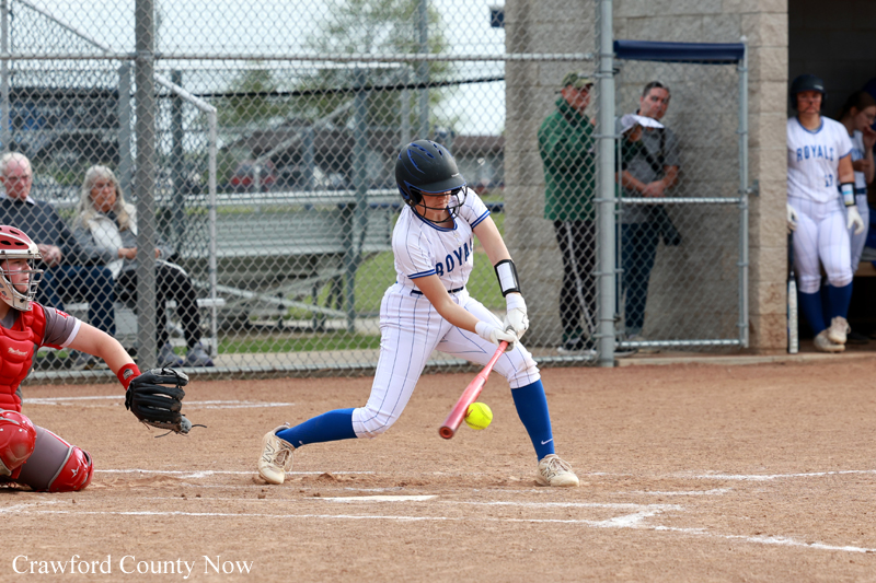 Female softball batter in white and blue swings at a pitched ball on a dirt field, with a red glove-wearing catcher crouched nearby.