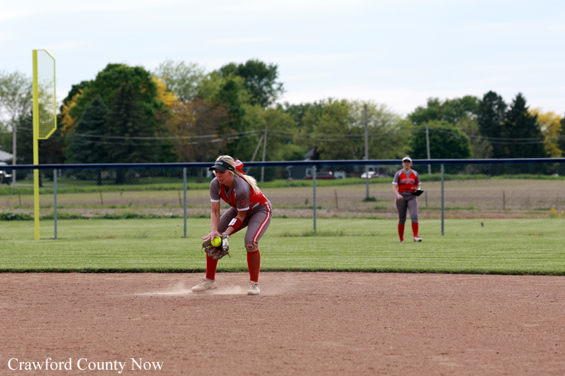 Female softball player in red uniform fields a ground ball on the infield as dust rises near her glove; another fielder stands in the outfield fence in the background.