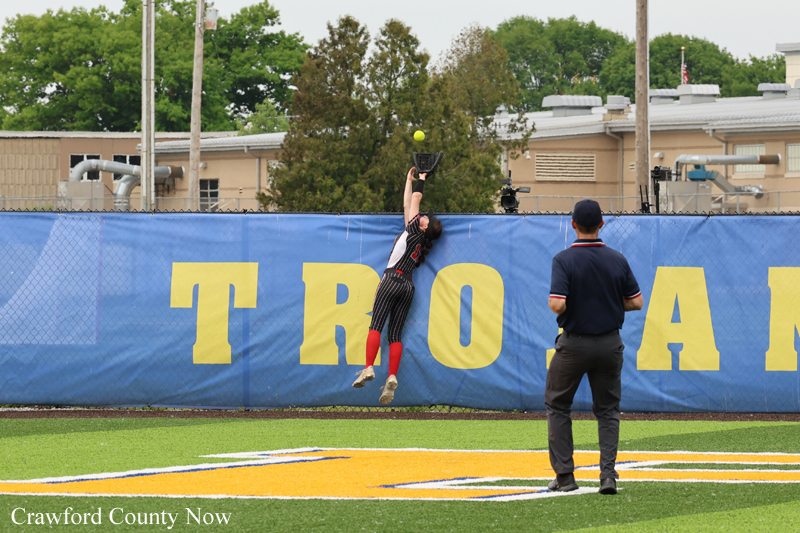 Softball/baseball fielder leaps to catch a ball against a blue outfield fence while an umpire watches nearby.