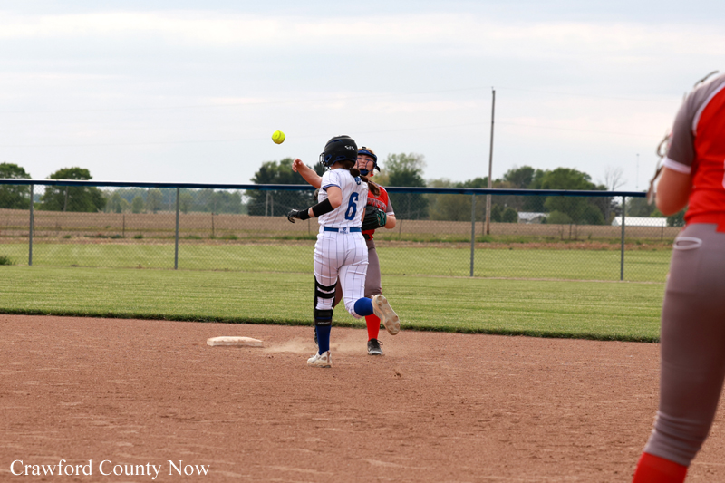 Softball player in a white uniform (#6) mid-throw near first base as a fielder in red pursues, ball in midair.