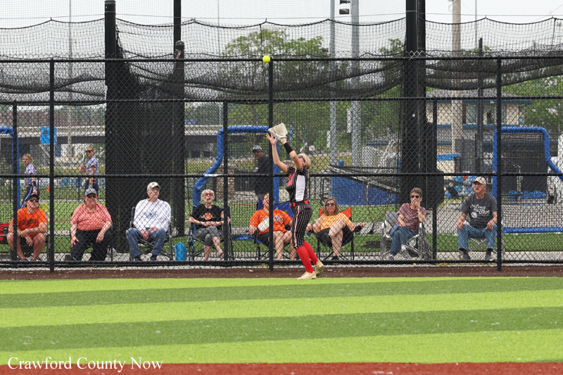 Female softball player in red socks reaches up to catch a ball near the outfield fence as spectators sit along the dugout behind the chain-link netting.