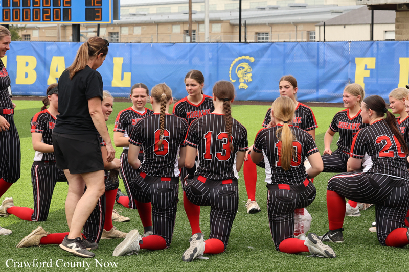 Girls' softball team in black pinstripe uniforms kneels in a huddle on a green field while a coach speaks to them. Behind them, a blue banner and scoreboard are visible across the field.