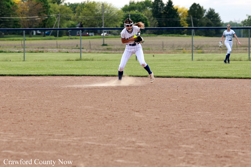 Female softball infielder in a white uniform fields a ground ball, dirt kicking up as she slides to grab it, with another fielder in the background on the grass.
