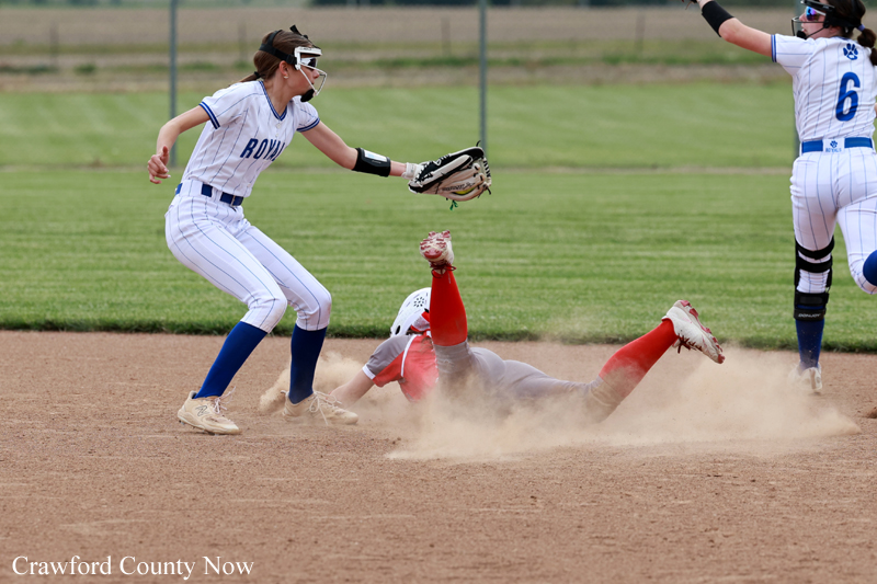 Softball player in white and blue reaches with glove as another runner slides into base, kicking up dust on the infield.