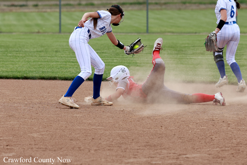 Female softball player slides into a base as a fielder reaches with a glove to tag, dust rising from the dirt infield.