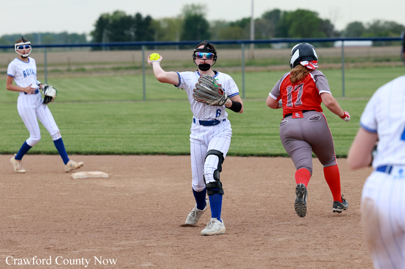 Softball game action: pitcher in a white uniform winds up to throw, glove and knee brace visible, as a runner in red jersey #11 sprinting past the infield. Other players stand on the dirt infield and grassy outfield.