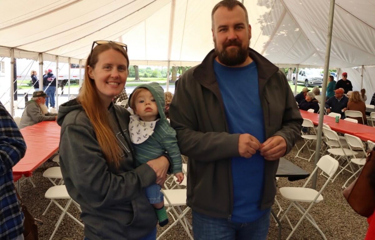 Mother with a baby in her arms and a bearded man standing beside under a tent at a community event.