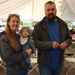 Mother with a baby in her arms and a bearded man standing beside under a tent at a community event.