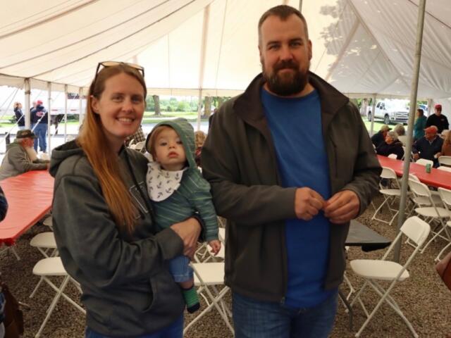 Mother with a baby in her arms and a bearded man standing beside under a tent at a community event.