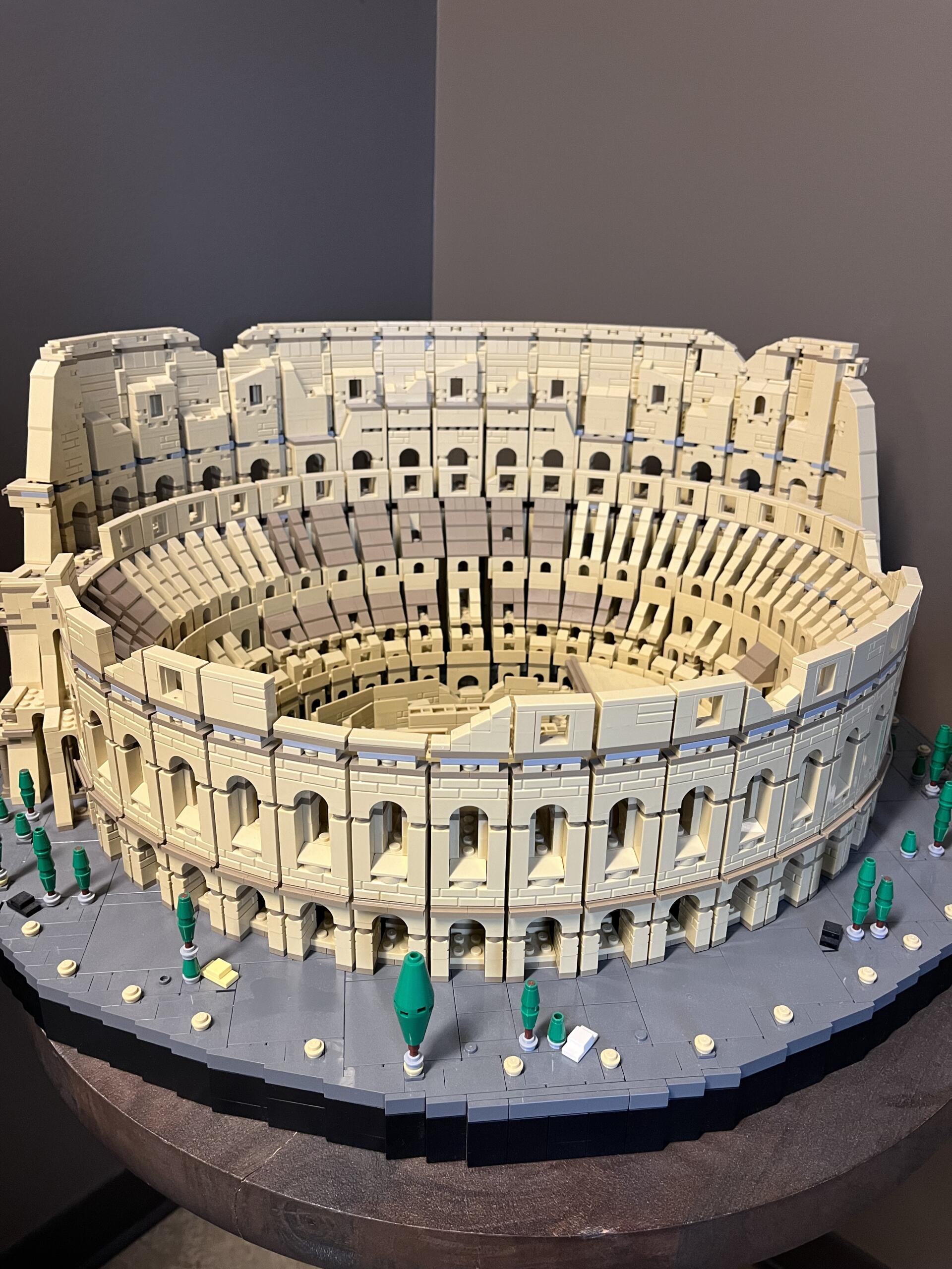 Large tan LEGO replica of the Colosseum, showing arched ground-level openings and tiered seating around a circular arena on a gray surface
