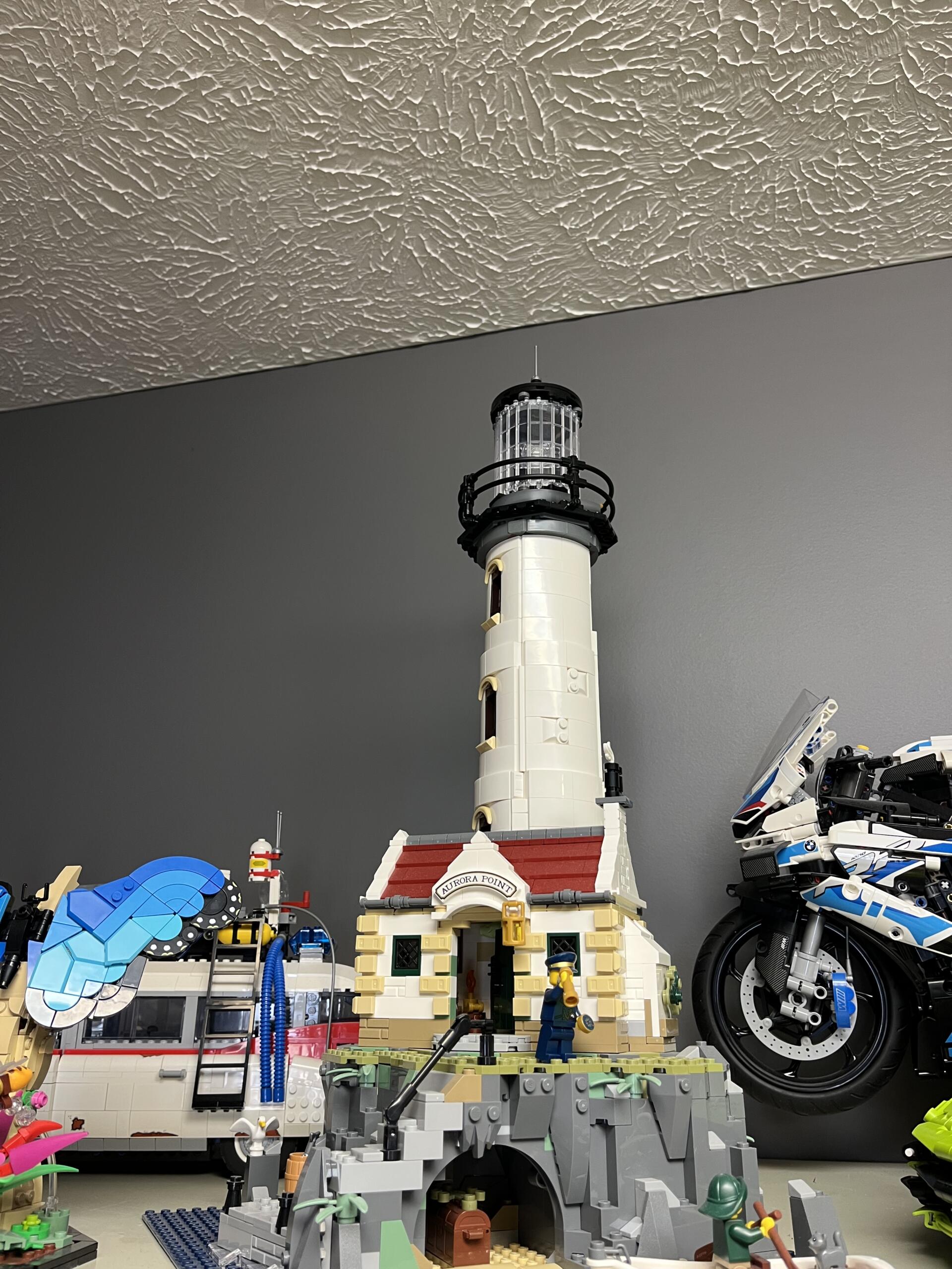LEGO lighthouse scene with a tall white tower on a rocky base and a red-roof building at the base, surrounded by various LEGO figures and accessories.