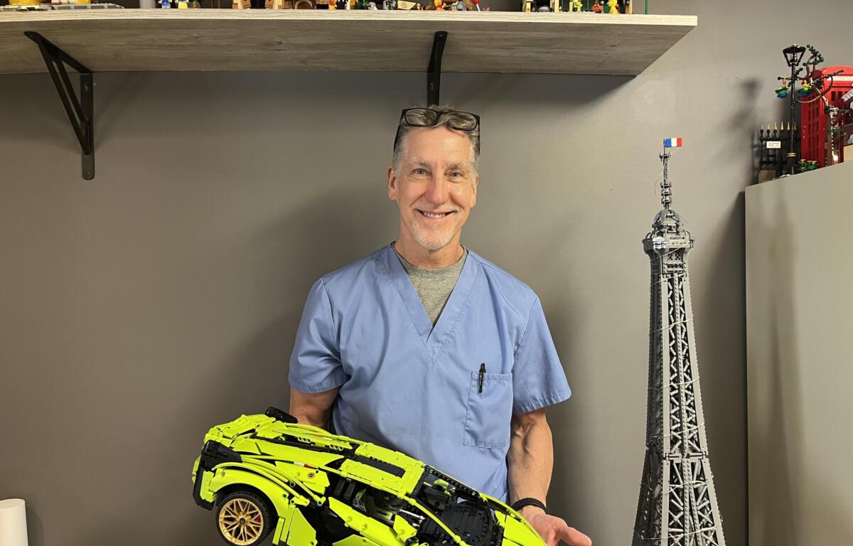 Smiling man in blue scrubs holds a neon yellow toy sports car in a room with toy shelves and a tall model tower in the background.