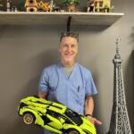 Smiling man in blue scrubs holds a neon yellow toy sports car in a room with toy shelves and a tall model tower in the background.