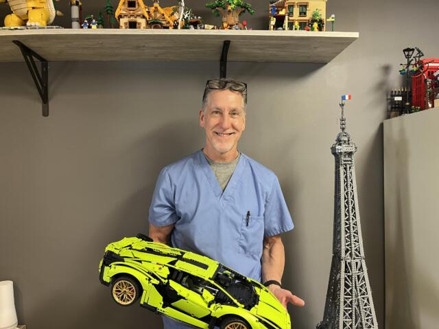 Smiling man in blue scrubs holds a neon yellow toy sports car in a room with toy shelves and a tall model tower in the background.