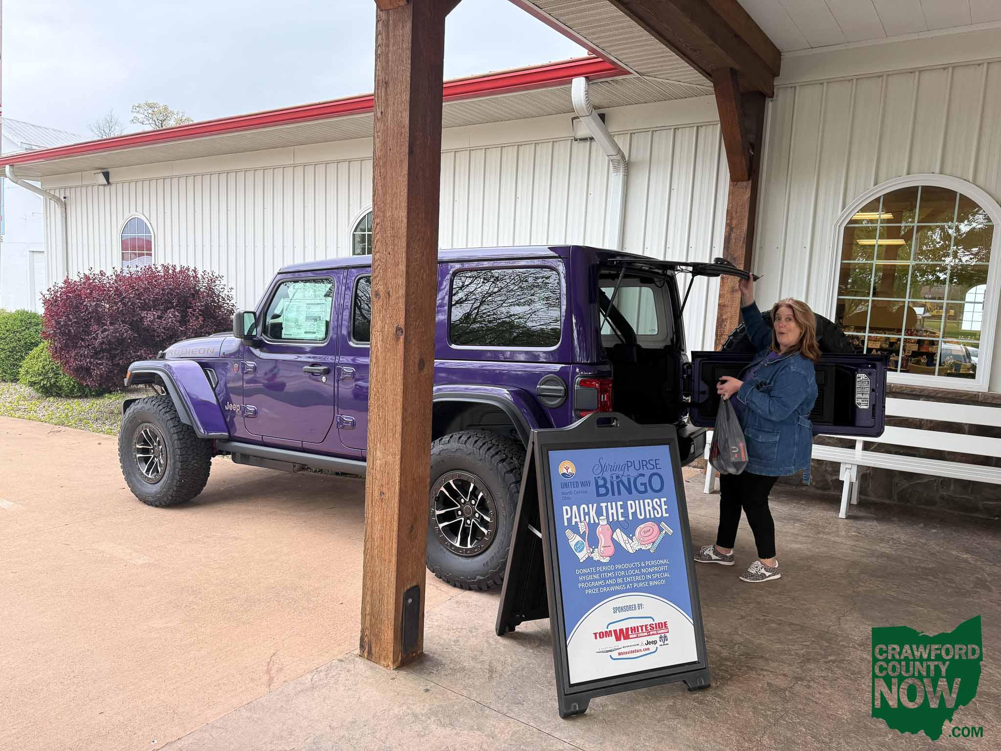Purple Jeep Wrangler parked under a wooden carport with a fundraising signboard nearby; a woman stands beside the open back of the vehicle.
