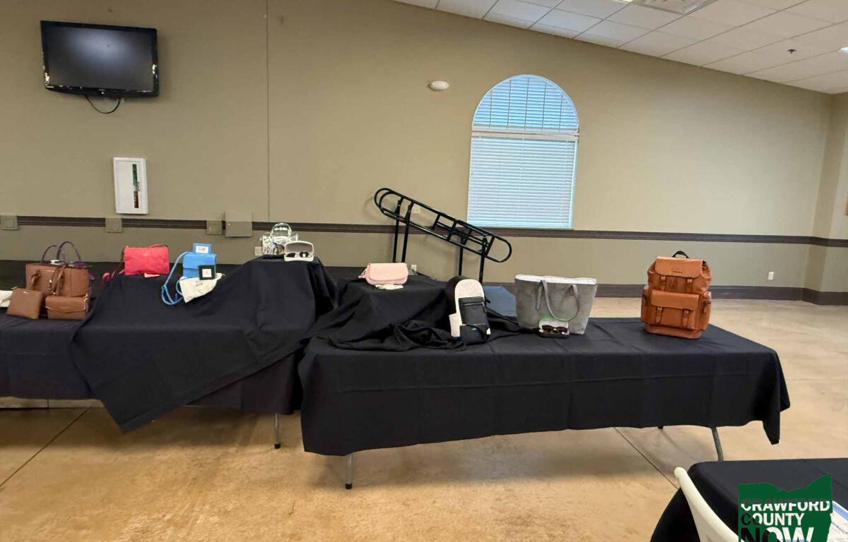 Tables covered with black cloth display bags and accessories in a room; wall-mounted TV, arched window, and Crawford County Now logo visible in the corner