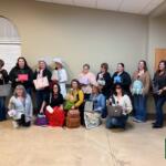 Group of women posing indoors with various handbags, some wearing hats, Crawford County Now logo visible in the bottom right corner