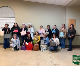 Group of women posing indoors with various handbags, some wearing hats, Crawford County Now logo visible in the bottom right corner