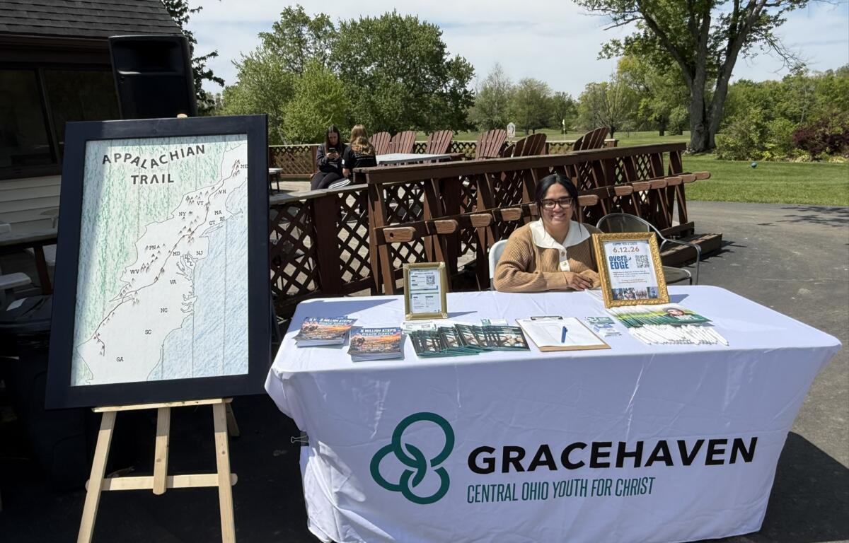 Outdoor information booth for Gracehaven with a framed Appalachian Trail map on an easel beside a table of brochures and a smiling woman sitting at the front.