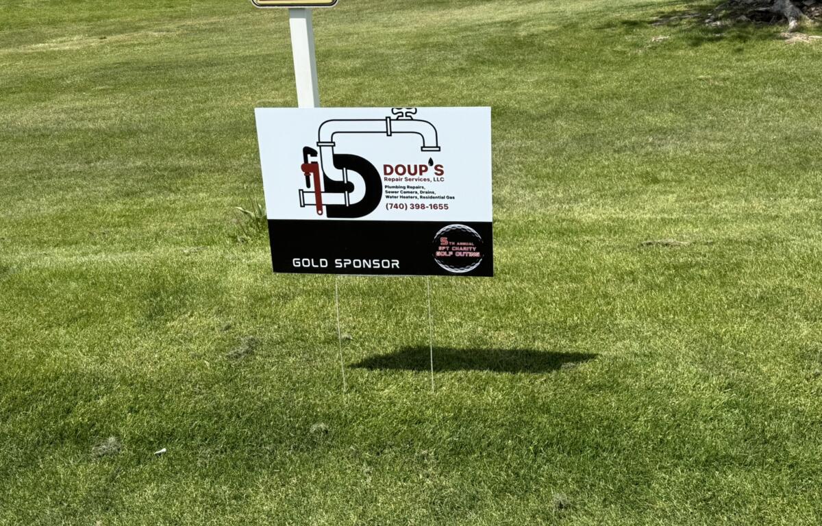 Golf course fairway with sponsor signs: a white sign showing a plumbing illustration for DOUP'S Plumbing as a Gold Sponsor, and a smaller Mansfield Brick & Supply sign above.