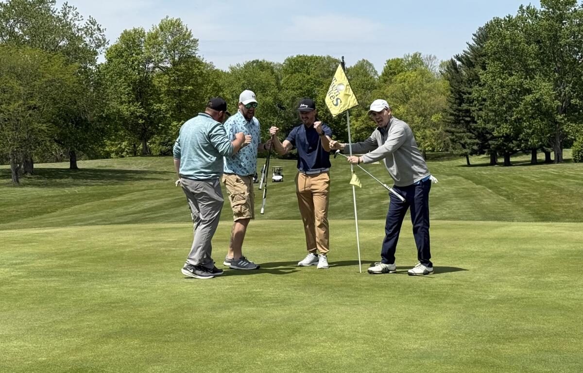Four men on a golf green celebrate together, fists raised near the hole with a yellow flag in the background.