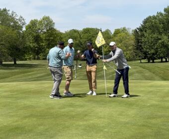 Four men on a golf green celebrate together, fists raised near the hole with a yellow flag in the background.