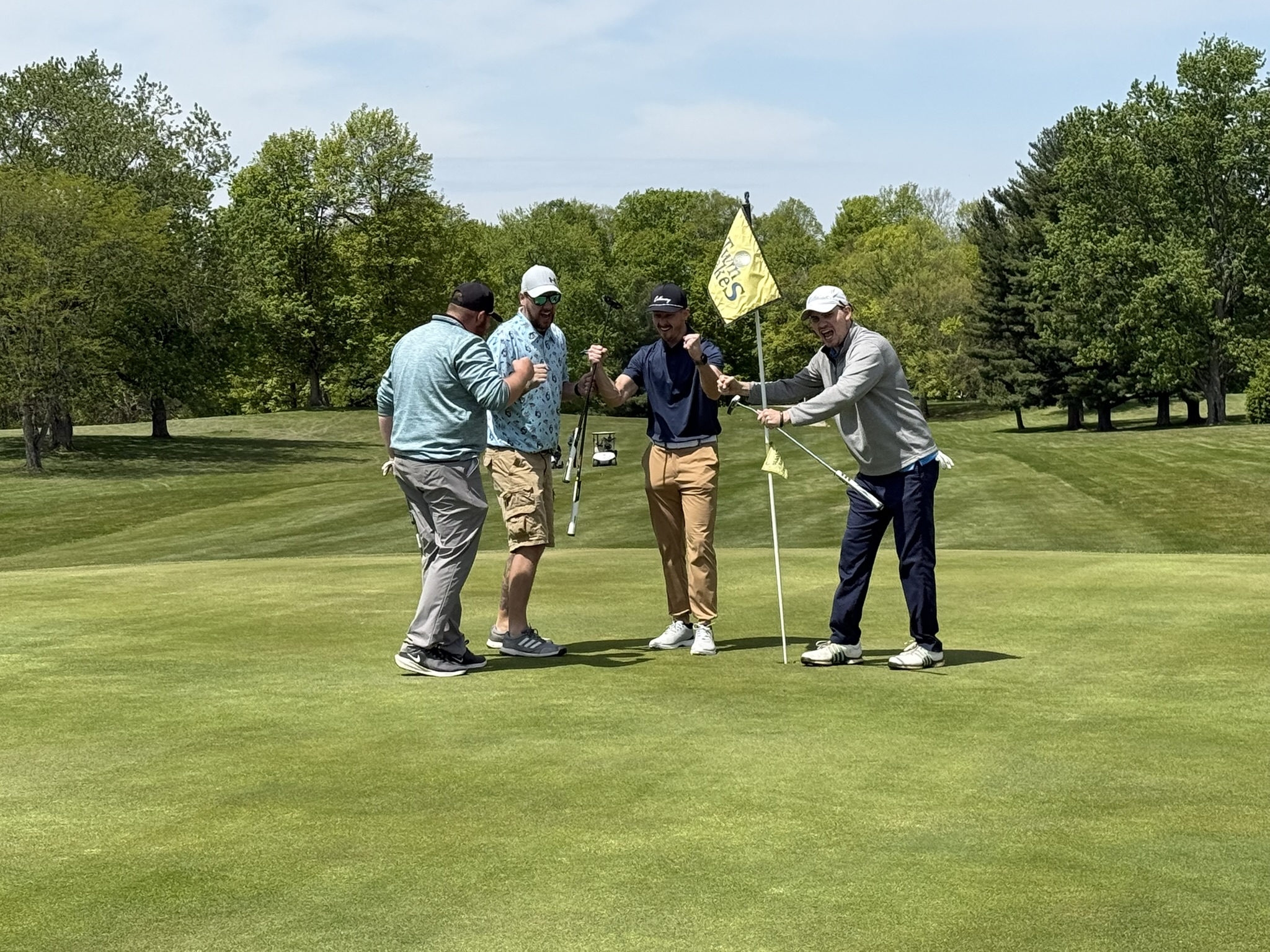 Four men on a golf green celebrate together, fists raised near the hole with a yellow flag in the background.