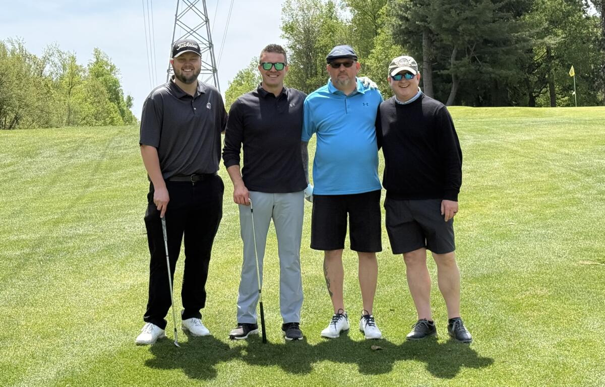 Four men posing together on a sunlit golf course, each with a golf club, smiling at the camera near a power line tower and trees in the background.
