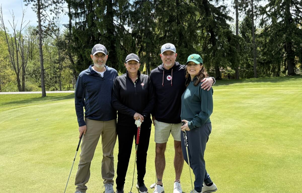 Four adults posing together on a golf course with clubs, smiling, trees in the background.