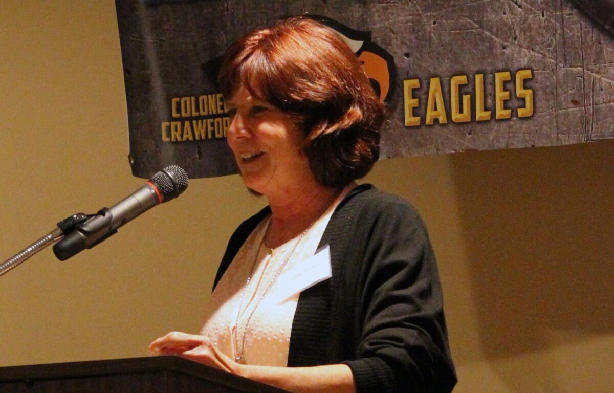 Woman speaks at a podium with a microphone, wearing a light blouse and dark cardigan, with an Eagles banner in the background.
