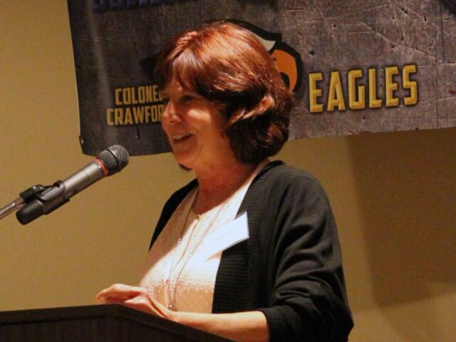 Woman speaks at a podium with a microphone, wearing a light blouse and dark cardigan, with an Eagles banner in the background.