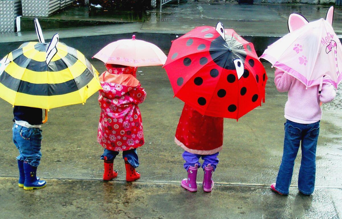 Children standing in a rainy street, each holding colorful cartoon-themed umbrellas seen from behind.