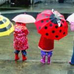 Children standing in a rainy street, each holding colorful cartoon-themed umbrellas seen from behind.