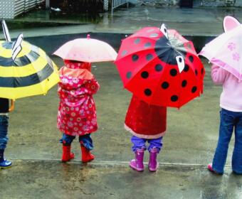 Children standing in a rainy street, each holding colorful cartoon-themed umbrellas seen from behind.