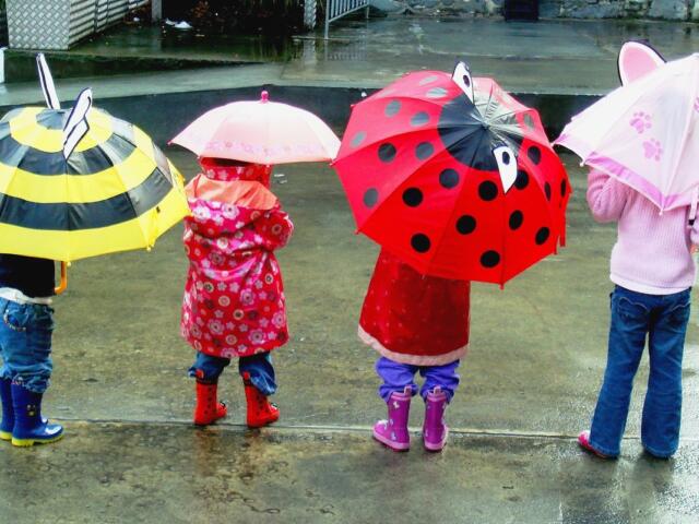 Children standing in a rainy street, each holding colorful cartoon-themed umbrellas seen from behind.
