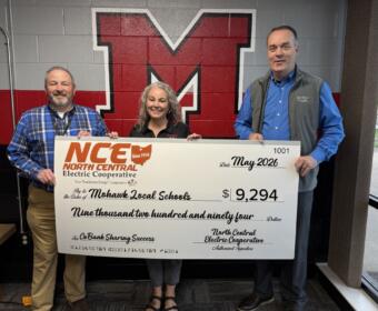 Three adults stand in front of a red and black wall mural holding a large ceremonial check from North Central Electric Cooperative for $9,294 to Mohawk Local Schools, dated May 2026.