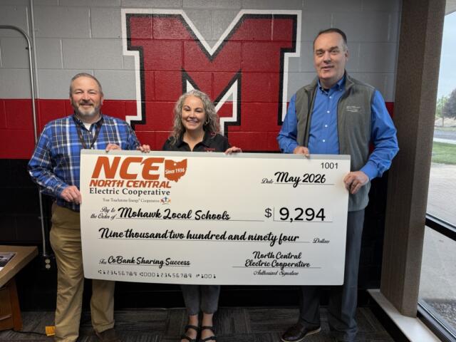 Three adults stand in front of a red and black wall mural holding a large ceremonial check from North Central Electric Cooperative for $9,294 to Mohawk Local Schools, dated May 2026.